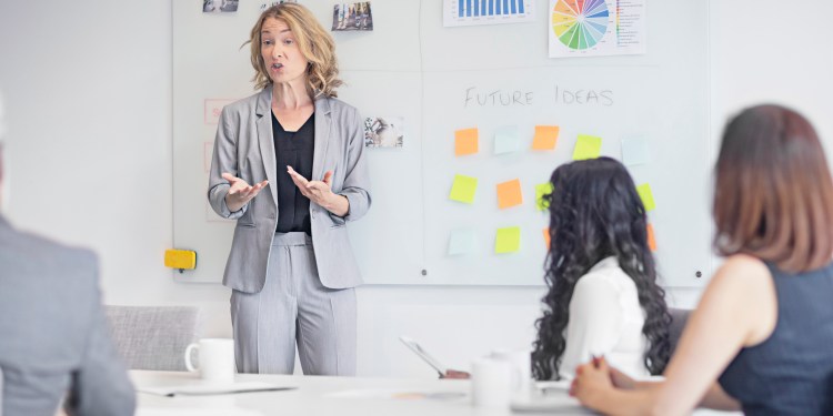 Female executive gesturing to white board while giving presentation to fellow professionals in office/workplace.