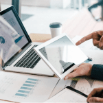 Person reviewing data analytics charts on a laptop and tablet at a desk with printed reports and a coffee cup.