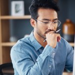 A man wearing glasses sits at a desk, thoughtfully looking at a computer monitor while holding a pen to his mouth, with shelves and office items in the background.